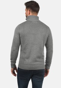 Grauer Strickpullover mit hohem Kragen, gerippten Bündchen und Saum. Verfügt über ein strukturiertes Muster und eine taillierte Passform, kombiniert mit dunklen Jeans.
