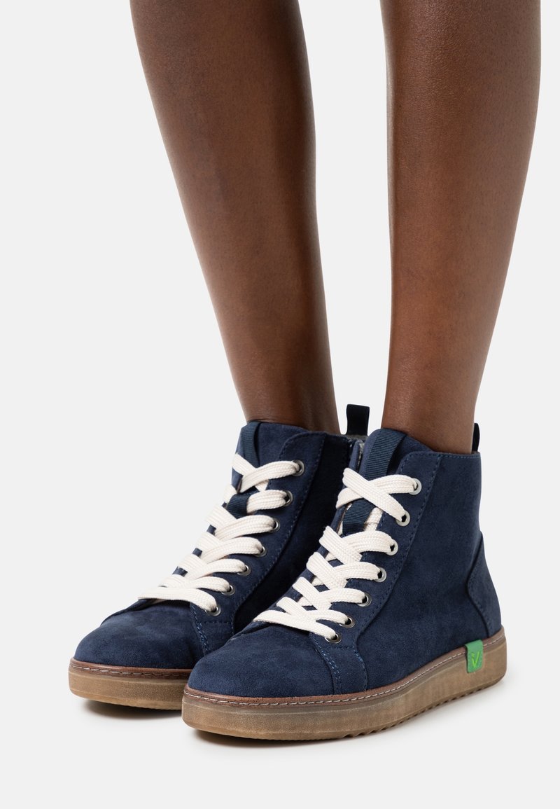 Bas des jambes d’une personne portant des baskets montantes en daim bleu marine avec des lacets blancs et des semelles marron clair sur un fond uni.