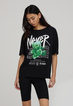 T-shirt oversize nera con grafica di un orsacchiotto verde e testo in grassetto, abbinata a shorts da bici neri. Il materiale sembra morbido e informale.