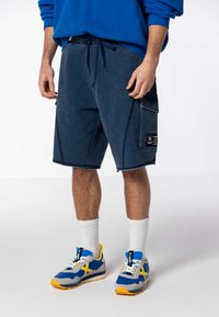 Shorts en denim bleu foncé, dotés d'une taille à cordon de serrage, de poches et d'une patch logo ; associés à des baskets multicolores et des chaussettes blanches.