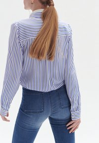 Camicia a righe con linee verticali blu e bianche, abbinata a jeans fitted blu scuro. Tessuto liscio e design con colletto classico.