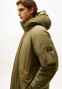 Parka d'hiver rembourrée vert olive avec une grande capuche, des poches zippées et un écusson logo sur la manche. Texture lisse et design utilitaire.