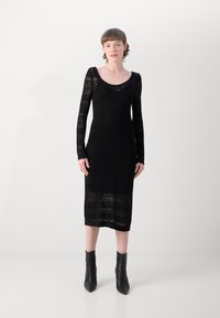 Abito in maglia nera con maniche lunghe, scollatura a girocollo e motivi testurizzati. Lunghezza a metà polpaccio. Abbinato a stivali neri alla caviglia.