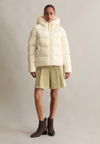 Cremet Puff-Jacke mit Kapuze und Reißverschluss, kombiniert mit einem grün gerippten Faltenrock und braunen Stiefeletten. Glatte Textur und lässige Passform.