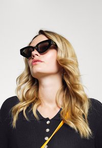 Óculos de sol pretos em formato de olho de gato com armações grossas, lentes escuras e detalhes em dourado nas hastes. Combinados com um top preto canelado e uma alça amarela.