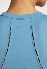 Vaaleansininen urheilupaita, jossa on pyöreä pääntie ja halkiot takana. Hienovarainen harmaa Nike-logo on ylhäällä. Sujuva ja joustava materiaali.