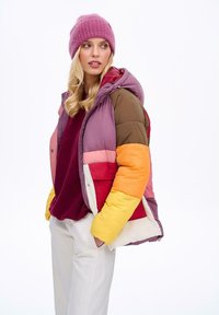 Prošívaná bunda s kapucí, s barevným blokovým vzorem v barvách fialová, hnědá, oranžová, červená a bílá. Nositelná přes bordó top.