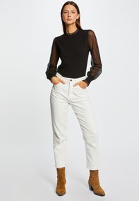 Maglione nero con dettagli in pizzo sulle maniche, abbinato a jeans bianchi a vita alta. Stivali alla caviglia marroni con tacco a blocco completano l'outfit.