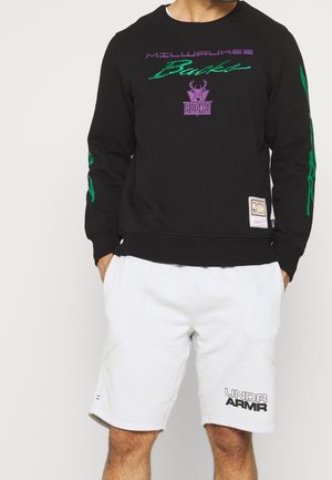 Sweatshirt noir avec des graphiques Milwaukee Bucks verts et violets, col rond et poignets côtelés, associé à un short gris clair avec un logo.