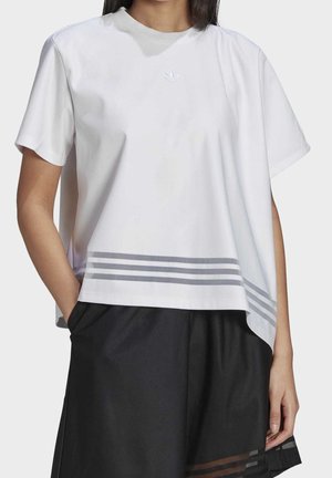 Persoon draagt een los wit T-shirt met een klein logo op de borst en drie horizontale grijze strepen nabij de zoom, gecombineerd met een zwarte broek.