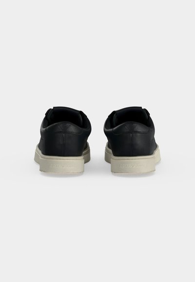 Paire de baskets basses noires avec semelles blanches vues de dos sur un fond clair uni.