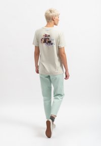 Lichtbeige t-shirt met een kleurrafische opdruk op de achterkant, gecombineerd met pastelgroene joggers. Zacht katoen, relaxed fit en korte mouwen.