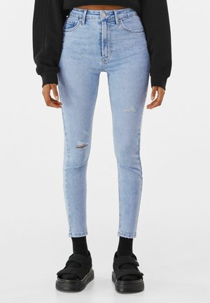 Personne portant un jean skinny bleu clair déchiré avec des chaussettes noires et des sandales noires à bout ouvert, debout devant un fond blanc.