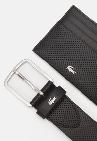 Curea din piele neagră perforată cu o cataramă din metal argintiu ce prezintă logo-ul Lacoste. Include un portofel de carduri negru asortat, cu textură netedă.