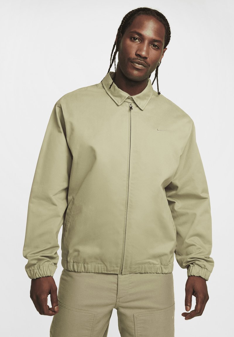 Veste bomber kaki avec un col, fermeture éclair à l'avant et poignets élastiques. Présente un logo Nike discret sur la poitrine. Texture lisse, design décontracté.