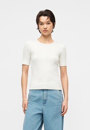 Jauns cilvēks ar īsiem tumšiem matiem, tērpies pieguļošā baltā īsroku kreklā un vaļīgās gaiši zilās džinsās, stāvot pret vienkāršu fona virsmu.