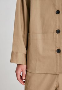Khaki napin kanssa löysä takki, jossa on rintatasku, pitkät hihat ja mustat napit. Kankaan pinta on sileä.