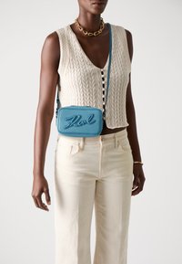 Geantă crossbody texturată de culoare albastră, cu litere embosate, având o curea subțire și o formă dreptunghiulară. Este purtată cu un top crem din tricot cu model de cabluri și pantaloni bej.