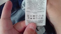 Uma etiqueta de cuidados com o tecido apresentando ícones de lavagem, secagem e engomagem, impressa em várias línguas, sobre um fundo texturizado branco.