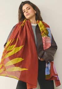 Kolorowy szal z cienkiej tkaniny na czerwonym tle, z wyraźnymi żółtymi liśćmi i motywami kwiatowymi w kolorze niebieskim i różowym, swobodnie ułożony.