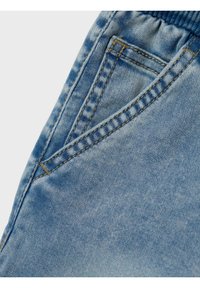 Tessuto di denim azzurro chiaro con una tasca frontale prominente, cuciture in contrasto e una finitura morbida e texturizzata. Dettaglio di vita elastica visibile.