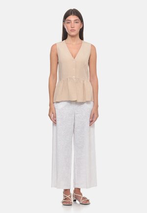 Donna in piedi, indossa un top beige senza maniche con bottoni e orlo a peplo, pantaloni bianchi larghi ricamati e sandali bianchi con cinturini.