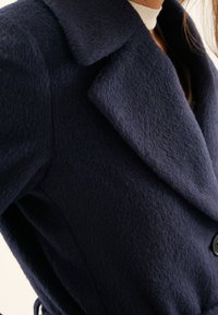 Námořnický vlněný kabát s texturovanou látkou, velkým klopovým límcem a zapínáním na knoflíky, s páskem v pase pro nastavitelnou velikost.