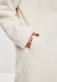 Faux-Fur-Mantel in Cremefarbe, mit strukturiertem Oberflächenmaterial, geradem Schnitt und einer Seitentasche mit sichtbarem Nahtdetail.