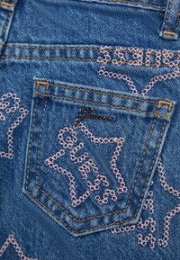 Kieszeń z dżinsu z różowym, haftowanym logo "GUESS" i ozdobnymi przeszyciami. Struktura ciemnoniebieskiej tkaniny z żółtym kontrastowym przeszyciem. Kształt heksagonalny.