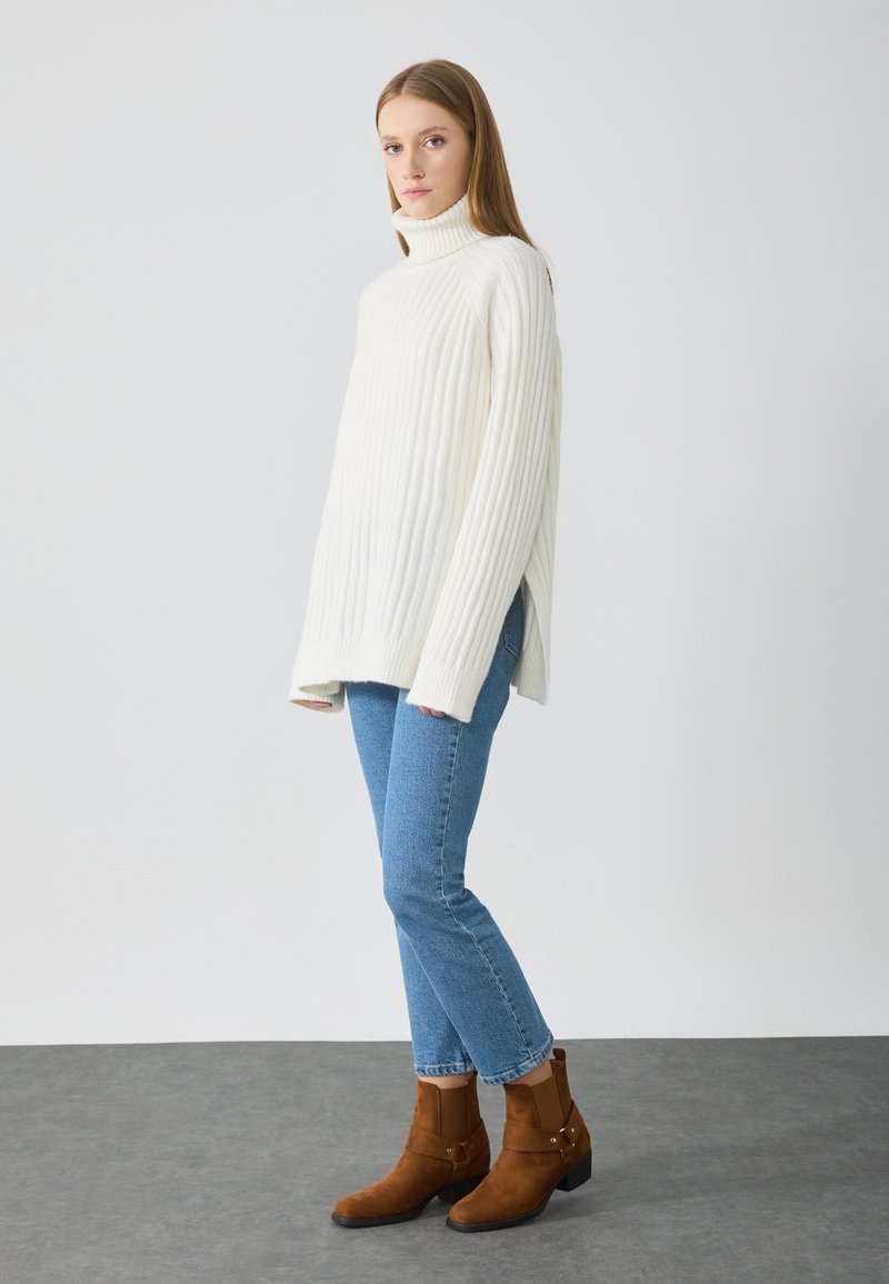 Suéter de cuello alto de canalé blanco grueso, de corte holgado, usado encima de jeans rectos azul claro y botines de ante marrón con tacón pequeño.