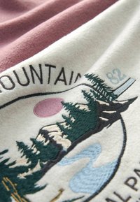 Koc polarowy w kolorze kremowym z różową sekcją. Posiada haftowany wzór drzew, gór i rzeki w obrębie okrągłej grafiki.