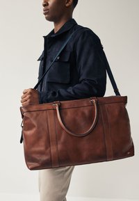 Brun lædertaske med tekstureret overflade, to robuste håndtag og en aftagelig skulderrem; har syninger som detaljer.