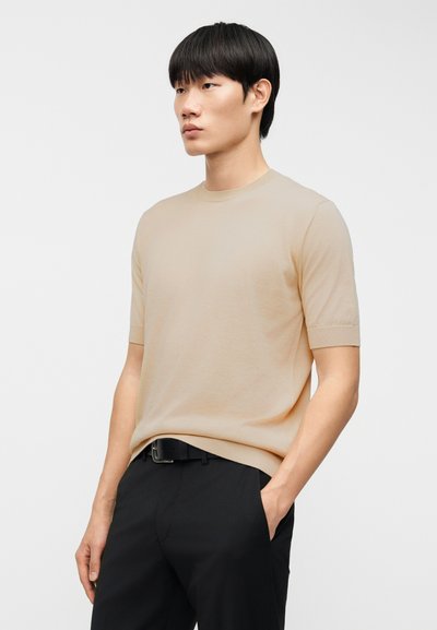 Μπεζ κοντομάνικη πλεκτή μπλούζα με στρογγυλή λαιμόκοψη, ομαλή υφή και ελαφρώς εφαρμοστή γραμμή, φορεμένη με μαύρο παντελόνι και ζώνη.