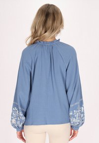 Blusa azzurra chiara con maniche lunghe e voluminose. Presenta un colletto arricciato e motivi floreali ricamati sui polsini delle maniche. Tessuto texture.