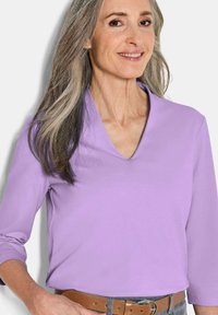 V-Ausschnitt lila Oberteil mit Dreiviertelärmeln, glatte Textur, lässige Passform, kombiniert mit einem braunen Gürtel über grauen Hosen.