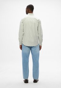 Μακρυμάνικη, ανοιχτοπράσινη, ριγέ πουκάμισο με κουμπωτές μανσέτες, φορεμένο με ανοιχτό μπλε τζιν και μαύρα παπούτσια, απεικονιζόμενο από πίσω.