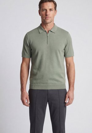 Mann trägt ein hellgrünes Polohemd mit kurzen Ärmeln und Frontreißverschluss, kombiniert mit dunkelgrauer Hose, steht vor einem einfarbigen Hintergrund.