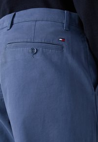 Kék pamut chino nadrág sima felülettel, hátoldalon gombos zsebbel és egy kis piros, fehér, és kék hímzett logóval.