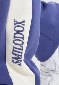 Sudadera morada con franjas laterales blancas. Presenta "SMILODON" bordado en azul en la manga izquierda. Tejido suave, corte deportivo.