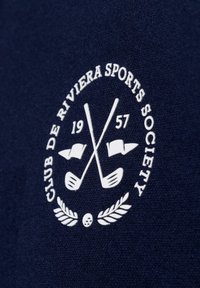 Dunkelblauem Stoff mit einem weißen gestickten Logo, das gekreuzte Golfschläger, Flaggen, den Text "Club de Riviera Sports Society" und Lorbeer-Akzente zeigt.