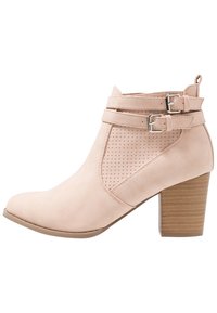 Bottine en faux suède rose clair avec accents de boucles doubles, design perforé sur le côté et talon en bois empilé. Texture lisse.