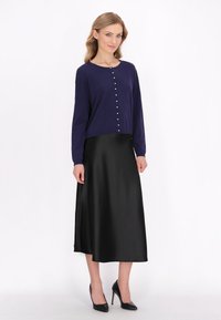 Cardigan lung cu mânecă lungă de culoare bleumarin, cu nasturi din perle, asociat cu o fustă midi din satin negru și pantofi cu toc înalt negri. Texturi netede în întreaga ținută.
