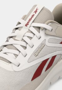 Zapatillas Reebok con parte superior de malla en un beige claro, acentos blancos, branding rojo y superposiciones texturizadas en los costados.