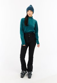 Błękitna, długą rękawą bluzka z zamkiem i prążkowaną teksturą, czarne, wysokie spodnie narciarskie z guzikami, szare buty narciarskie oraz robiona na drutach czapka. 