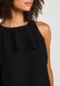 Femme aux cheveux bouclés portant un haut noir sans manches avec un détail de volant superposé et un fin collier en or sur un fond neutre.