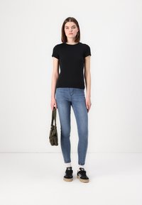 Tricou negru cu mânecă scurtă, cu un design ajustat, asortat cu blugi skinny albaștri. Modelul ține în mână o geantă verde măsliniu, purtând adidași negri cu talpă bej.