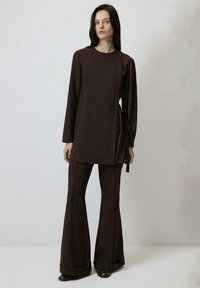Tunică lungă maro cu mâneci lungi, cu legătură pe lateral, purtată cu pantaloni evazați asortați. Material neted și design minimalist fără modele.