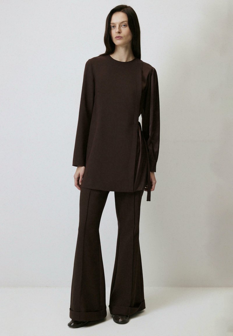 Tunică lungă maro cu mâneci lungi, cu legătură pe lateral, purtată cu pantaloni evazați asortați. Material neted și design minimalist fără modele.