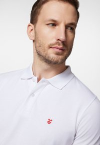 Biała koszulka polo wykonana z bawełny o teksturowanym wykończeniu. Posiada zapięcie na trzy guziki oraz małe haftowane czerwone logo na klatce piersiowej.