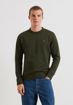 Young man wearing a dark green long-sleeve sweatshirt and blue jeans, standing with one hand in pocket against a plain light background.
